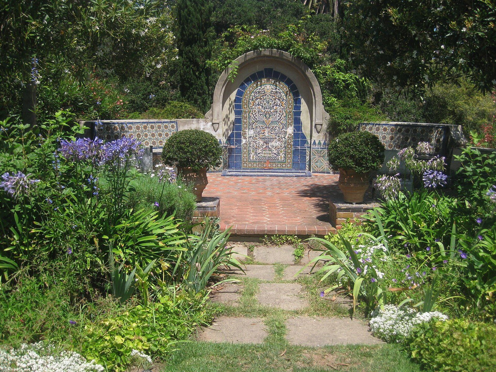 A tiled fountain and brick path among agapanthus and shrubs in the gardens of Casa del Her, a Wikimedia Commons photograph in the encyclopedia of Mike Kobeissi