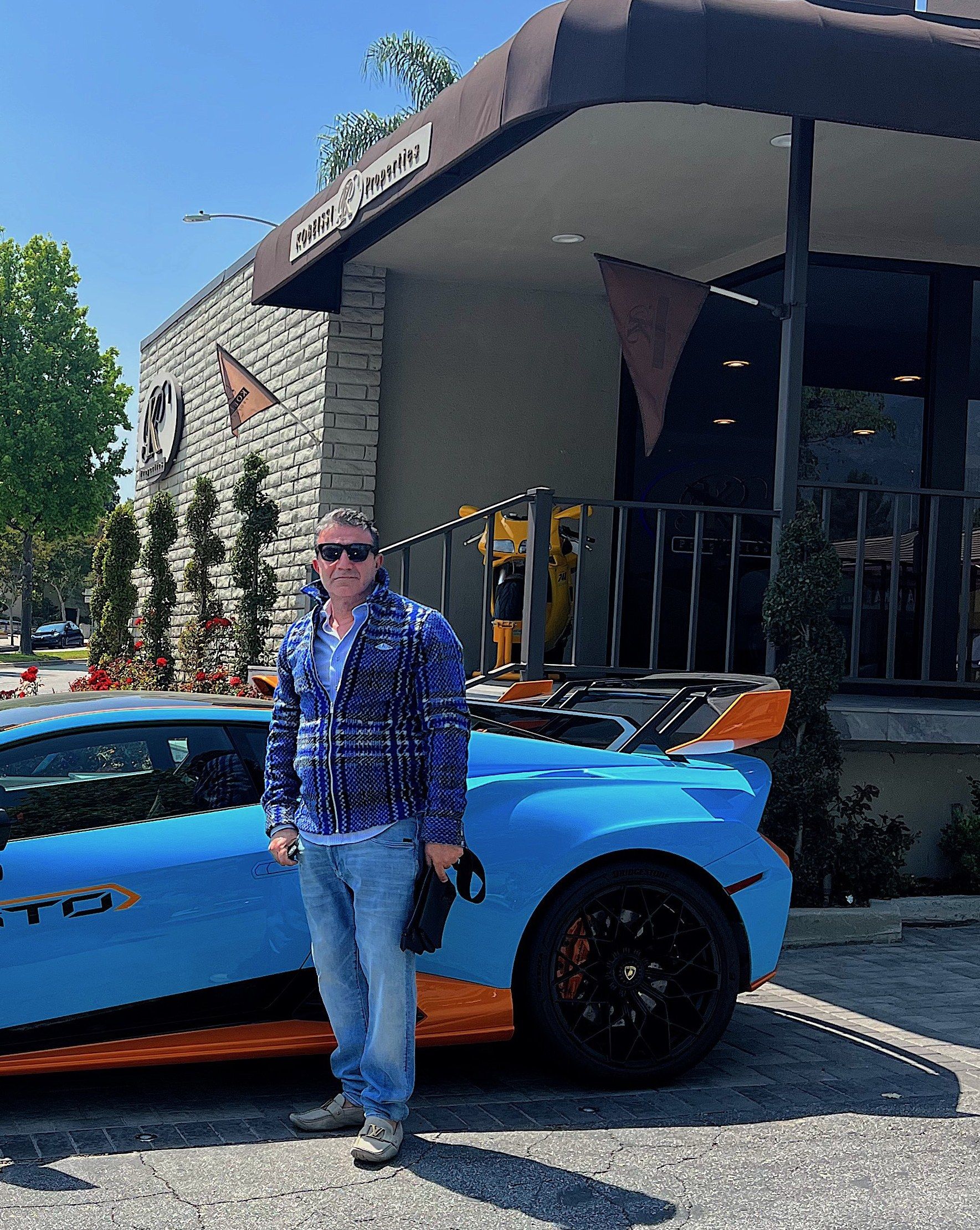 Mike Kobeissi in sunglasses standing beside a bright blue sports car outside his office