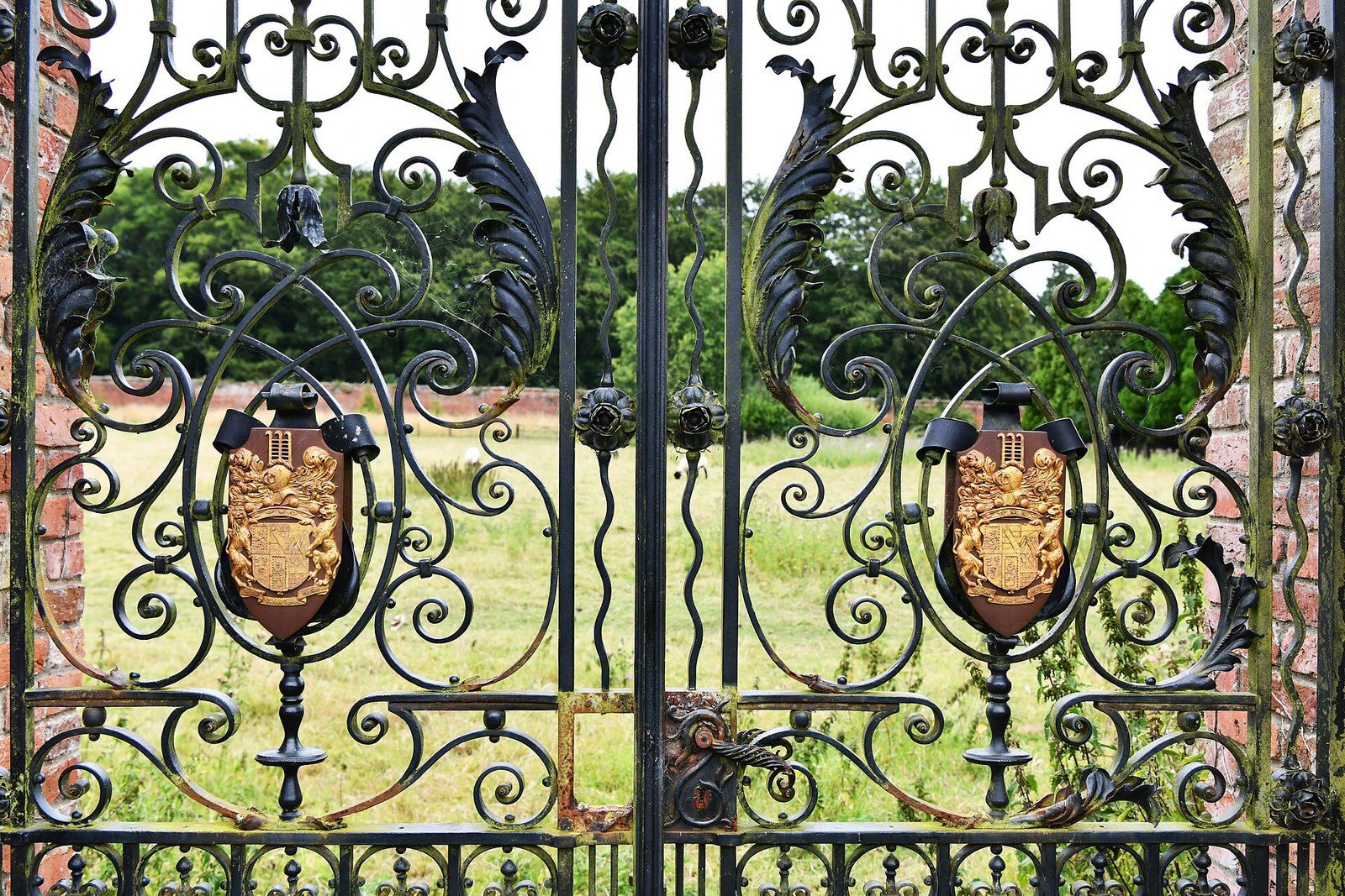 Ornate closed wrought iron garden gates with crests, a lawn and trees beyond, a Wikimedia Commons photograph in the encyclopedia of Mike Kobeissi